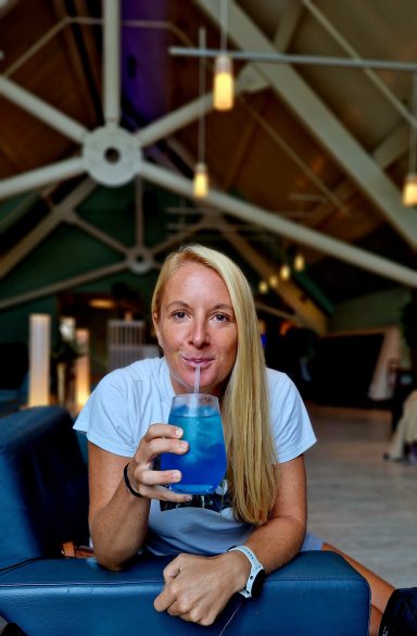 Junge Frau mit blonden Haaren trinkt einen blauen Cocktail in einem modernen Ambiente.