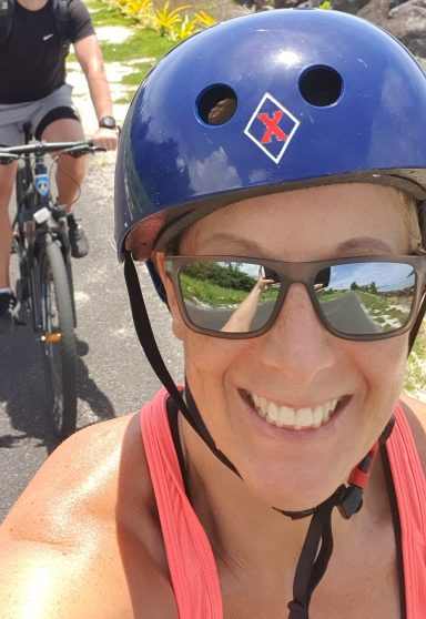 Frau mit Sonnenbrille und Helm lächelt, im Hintergrund ein Radfahrer auf einem Weg.