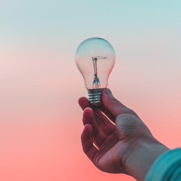 Eine Hand hält eine Glühbirne vor einem pastelgefärbten Himmel.