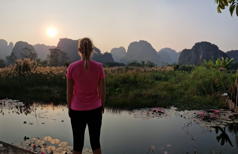 Person in rosa Shirt steht am Wasser und blickt auf eine Berglandschaft bei Sonnenuntergang.