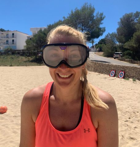 Lächelnde Frau mit großen Schwimmbrille am Strand, im Hintergrund sind Bäume und Gebäude.