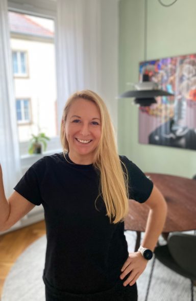 Lächelnde Frau mit blonden Haaren steht in einem hellen Raum neben einem Tisch.